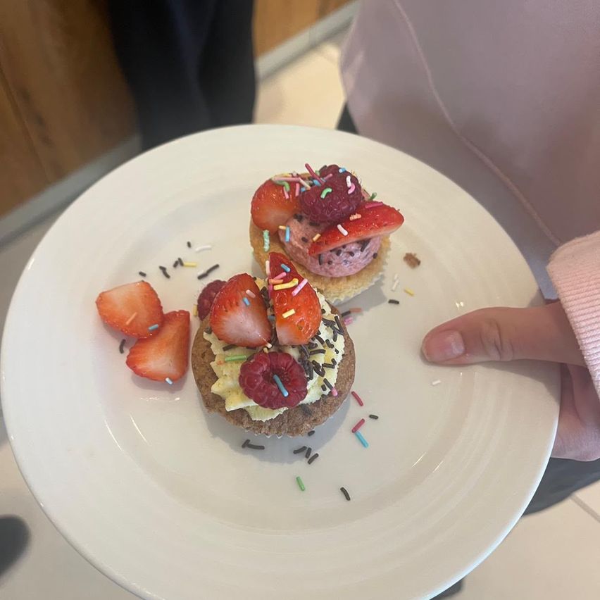 A white plate holds two cupcakes with strawberries, raspberries, and sprinkles. One is on the plate, and the other is held by a hand.
