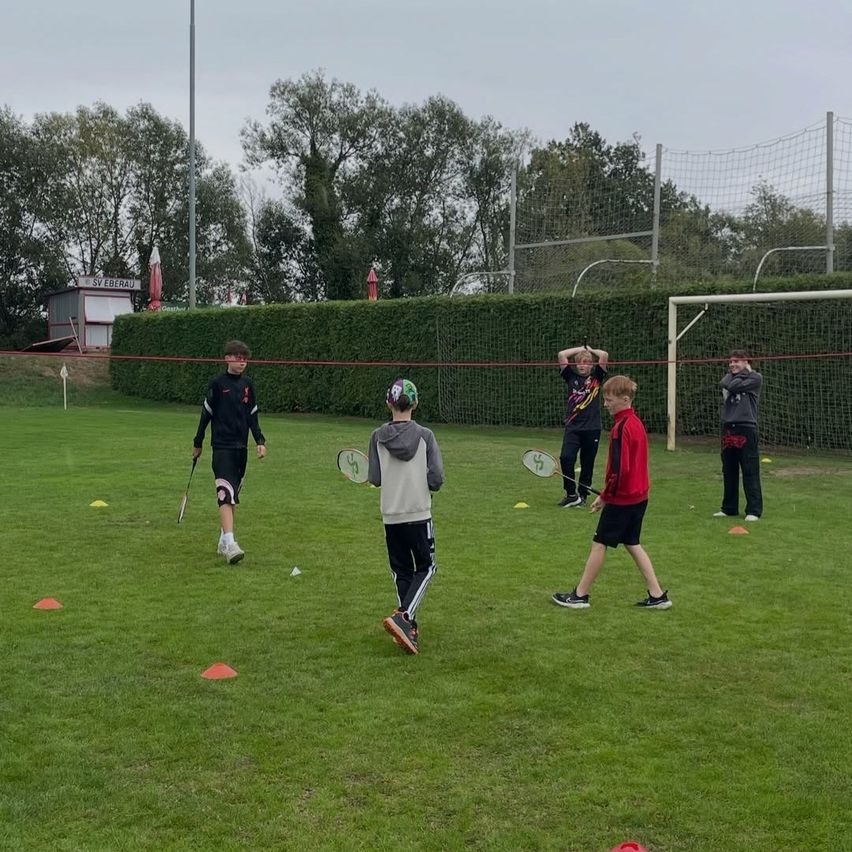 Fünf Jungen üben Tennis auf einem Rasenfeld mit orangefarbenen Hütchen und einem Netz im Hintergrund.