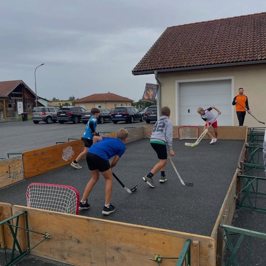 Fünf Kinder spielen Hockey auf einem improvisierten Spielfeld auf einem Parkplatz. Ein Mann beobachtet von einem Gebäude aus.
