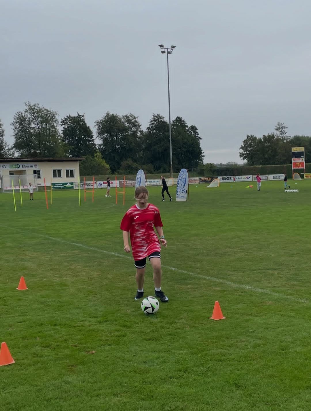 Ein junger Junge in einem roten Shirt übt das Kicken eines Fußballs auf einem Feld mit orangefarbenen Hütchen und anderen Spielern im Hintergrund.