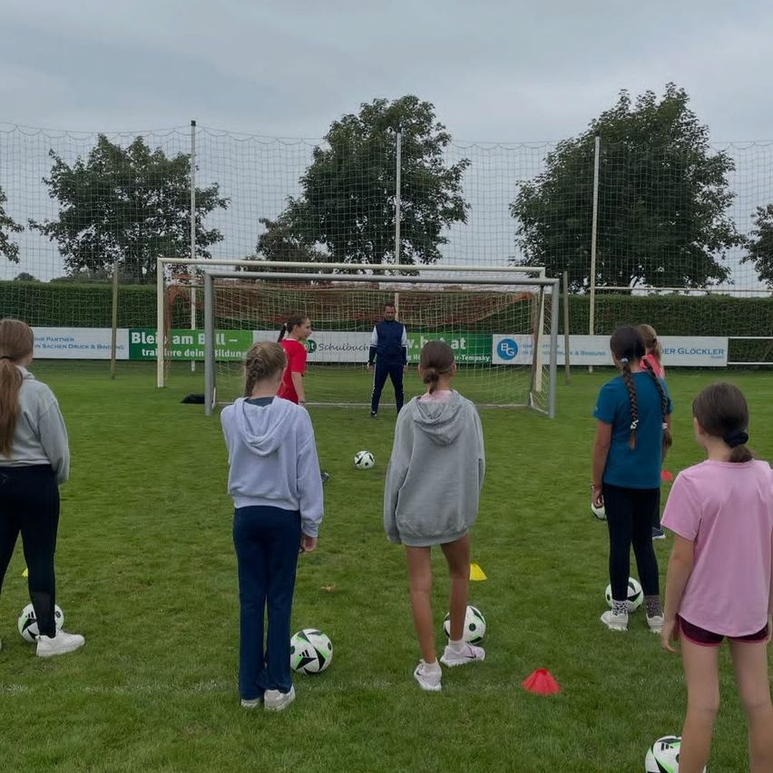 Eine Gruppe junger Mädchen trainiert Fußball auf einem Feld. Sie stehen vor einem Tor mit einem Mann in einem blauen Jackett. Es gibt Fußballbälle und Hütchen auf dem Rasen.