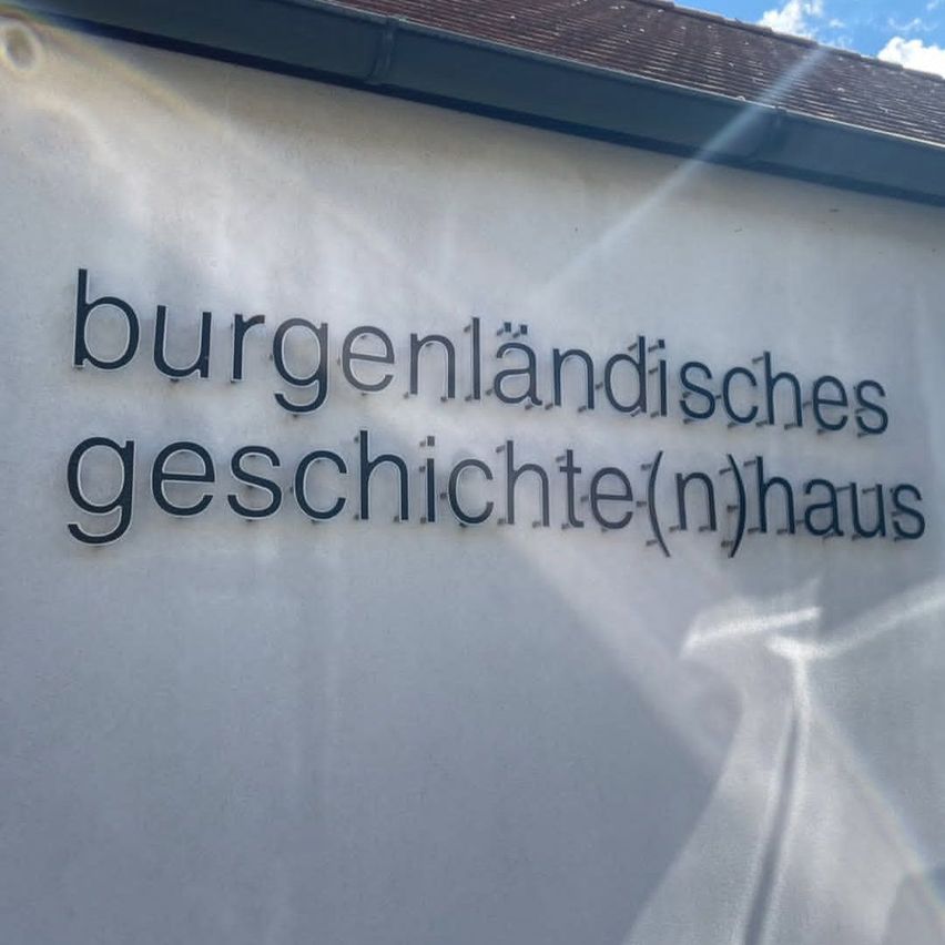 Eine weiße Wand mit einem Schild, das 'burgenlandisches geschichte(n)haus' in schwarzen Buchstaben steht. Das Schild ist an der Wand montiert und die Wand wird von Sonnenlicht beleuchtet.