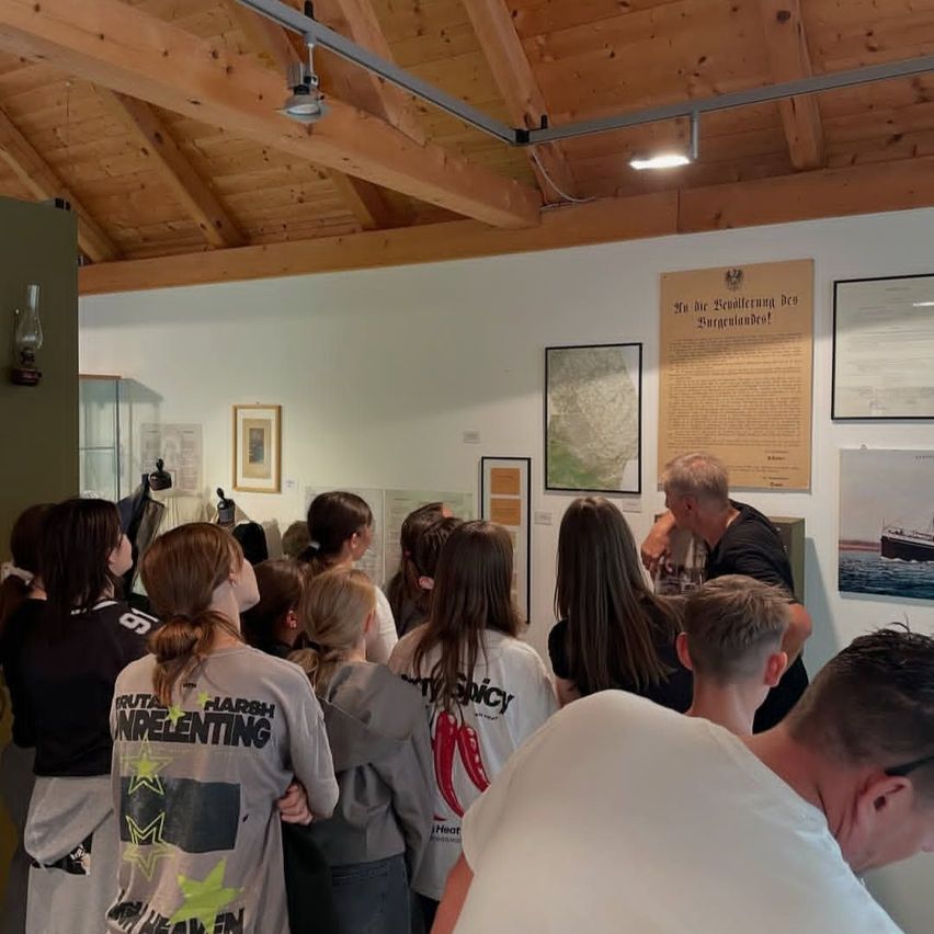 Eine Gruppe von Menschen versammelt sich in einem Raum mit Holzdecken und Wänden und betrachtet verschiedene Ausstellungen. Ein Mann spricht zu ihnen.