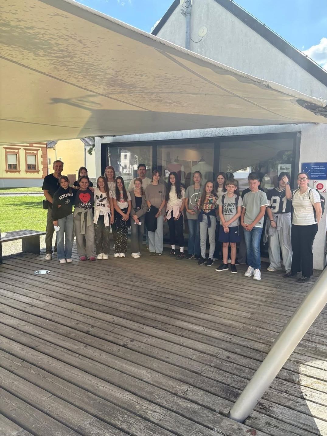 Eine Gruppe von Menschen, darunter Erwachsene und Kinder, posiert für ein Foto vor einem Gebäude. Sie stehen auf einer hölzernen Terrasse unter einem Vordach.