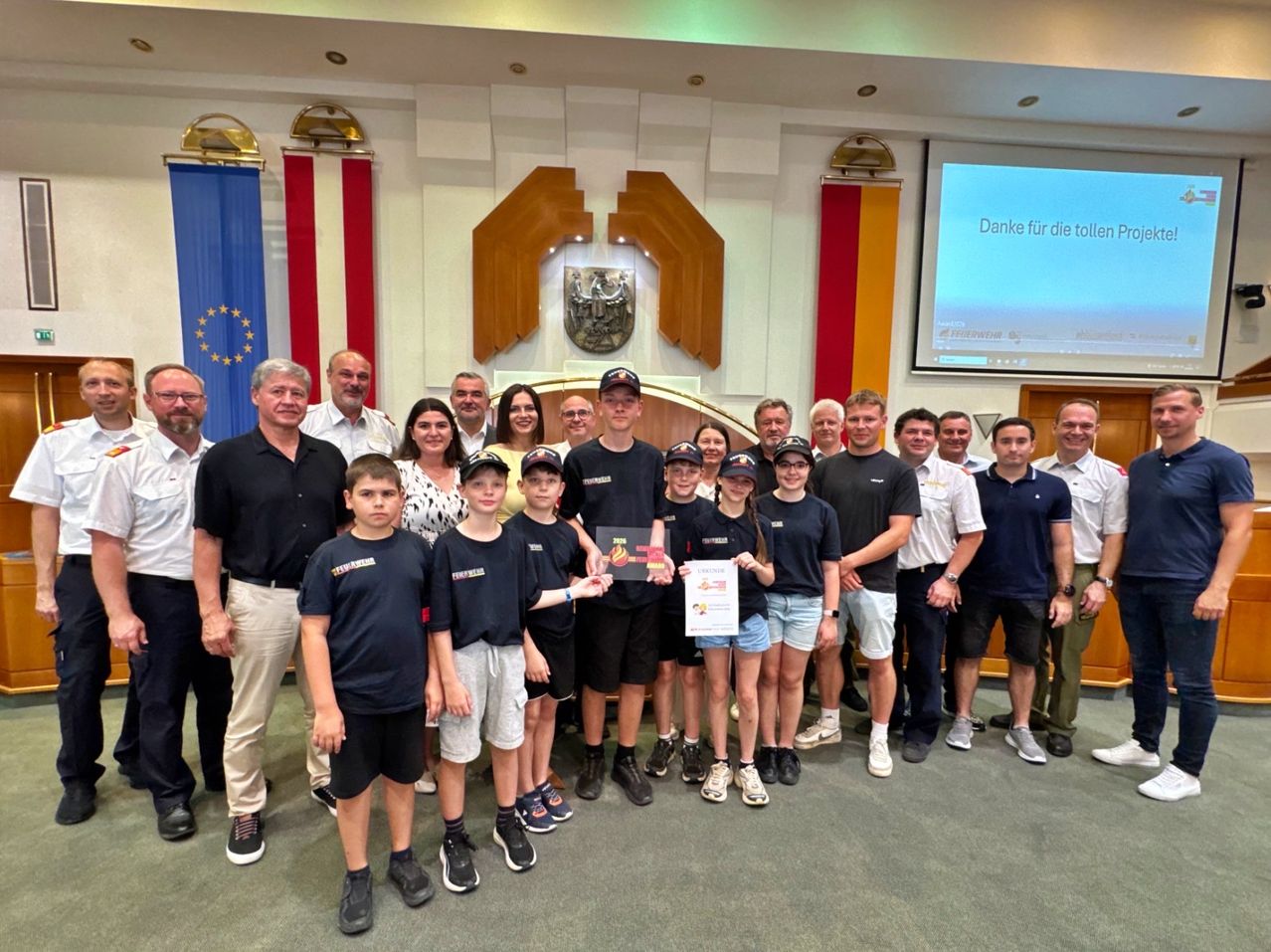 Eine Gruppe von Menschen, einige Erwachsene und einige Kinder, posiert für ein Foto in einem Konferenzraum. Die Kinder halten Urkunden in den Händen. Hinter ihnen befindet sich eine Holzwand mit Flaggen und einer Projektionsleinwand.