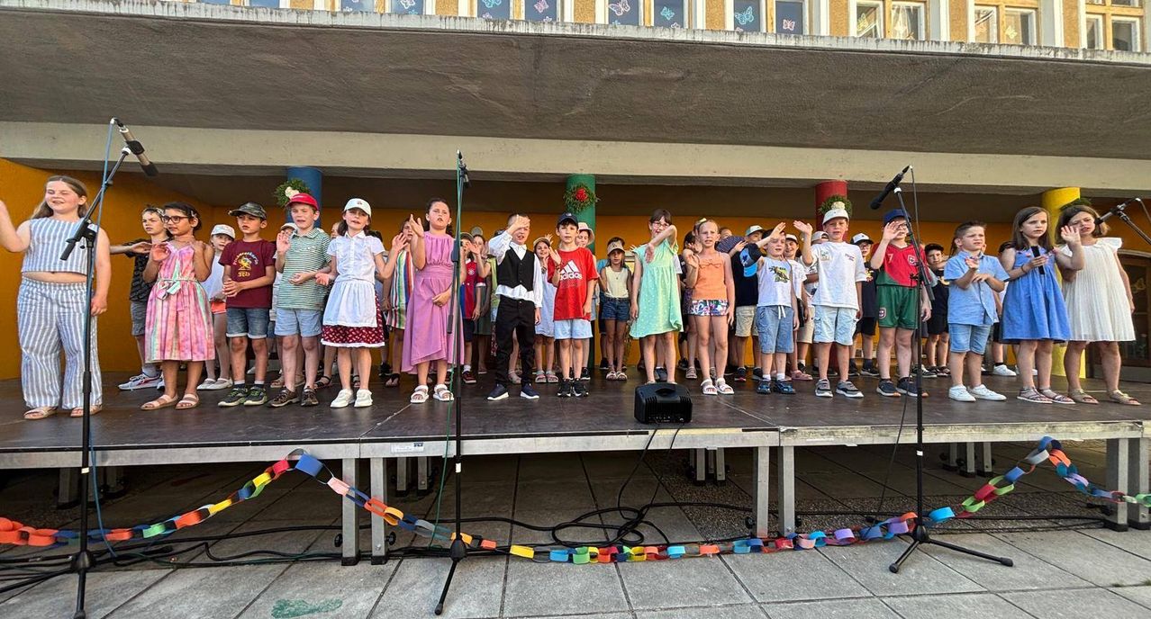 Eine Gruppe von Kindern tritt auf einer Bühne auf, mit Mikrofon, bunten Dekorationen und einem Hintergrund mit Schmetterlingsdesigns.