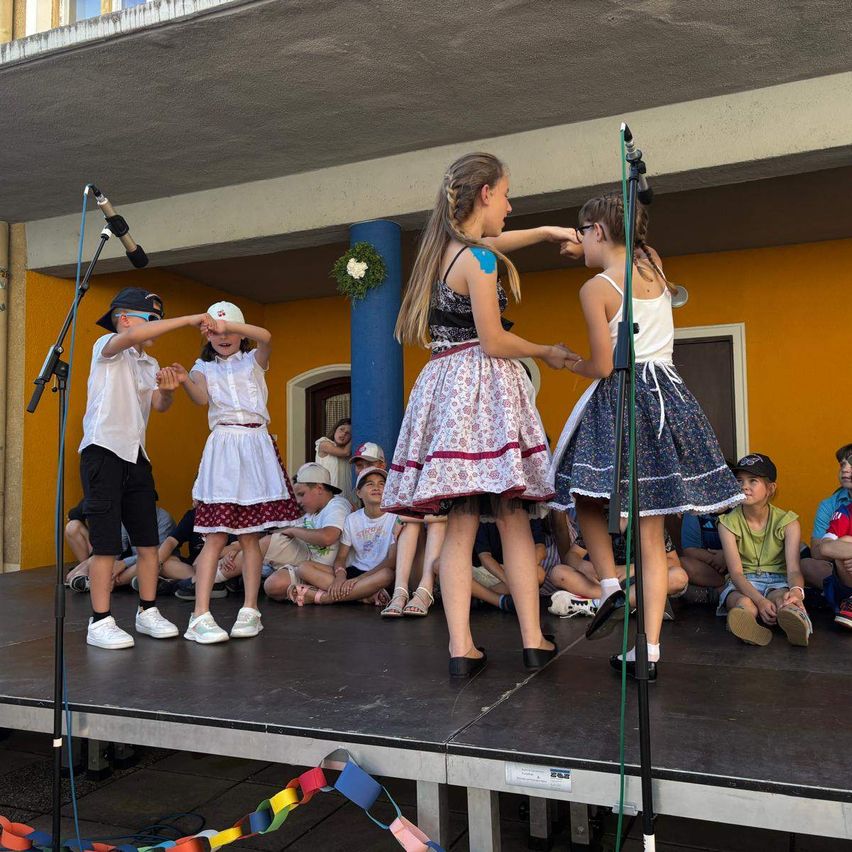 Eine Gruppe von Kindern in traditioneller Kleidung tritt mit zwei Mikrofonen auf der Bühne auf, während andere auf dem Boden dahinter sitzen.