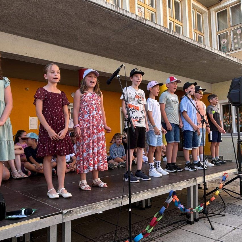 Eine Gruppe von Kindern singt auf der Bühne, sie tragen Hüte und Schuhe. Dahinter sitzt ein Publikum auf dem Boden. Ein Mikrofon und ein Lautsprecher sind auf der Bühne aufgebaut.