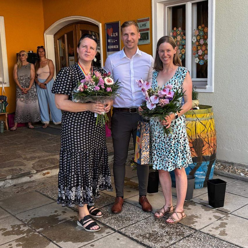 Drei Personen, zwei Frauen und ein Mann, stehen zusammen und halten Blumen. Sie scheinen ein Ereignis zu feiern. Hinter ihnen stehen zwei Frauen und ein Fass. Die Wand ist gelb gestrichen mit einem Schild und einem Fenster mit Blumendekoration.