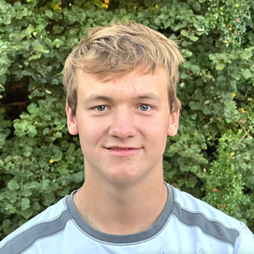 Ein junger Mann mit blonden Haaren und blauen Augen lächelt. Er trägt ein hellblaues und graues T-Shirt mit einem grauen Kragen. Im Hintergrund befinden sich dichte grüne Blätter.