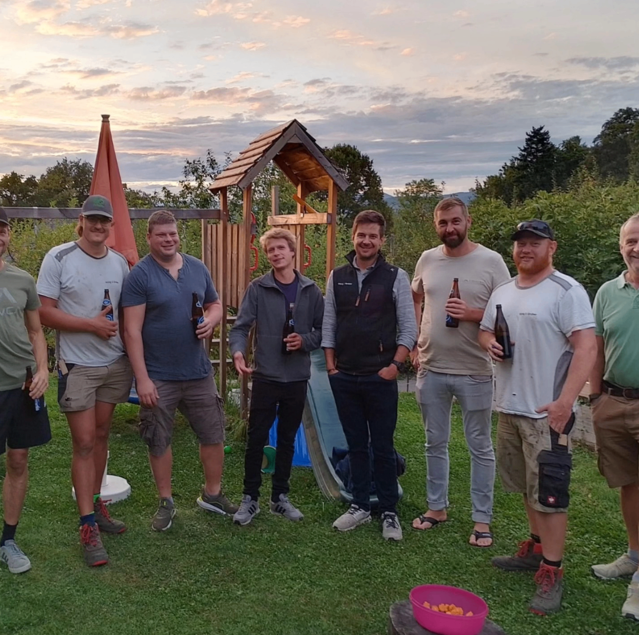 Eine Gruppe von Männern steht in einem Garten, lächelt und hält Flaschen. Sie sind lässig gekleidet, und einige tragen Mützen. Hinter ihnen ist eine kleine Holzstruktur und eine Rutsche zu sehen. Bäume und ein Zaun sind im Hintergrund.