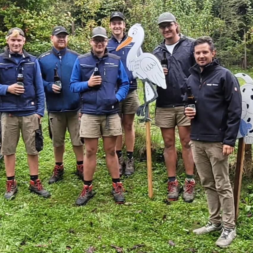 Eine Gruppe von sechs Männern auf einem Feld mit Flaschen in der Hand, einer steht hinter einer Ausschnitt-Stork. Bäume und Pflanzen im Hintergrund.