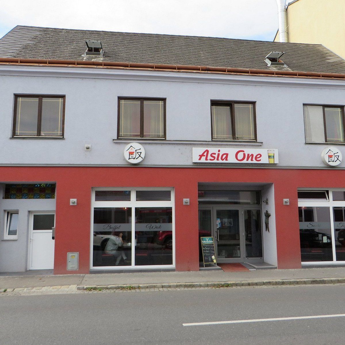 Die Vorderseite des Asia One Restaurants. Es hat eine rote Wand, Glasfenster und eine Glastür. Ein Menübrett steht vor dem Eingang. Drinnen sitzt eine Person an einem Tisch.