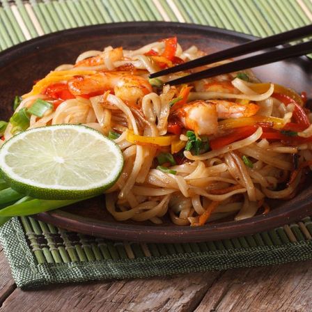 Eine Schüssel mit Garnelennudeln und Gemüse, dazu eine Limettenscheibe und Chopsticks auf einem Holztisch.