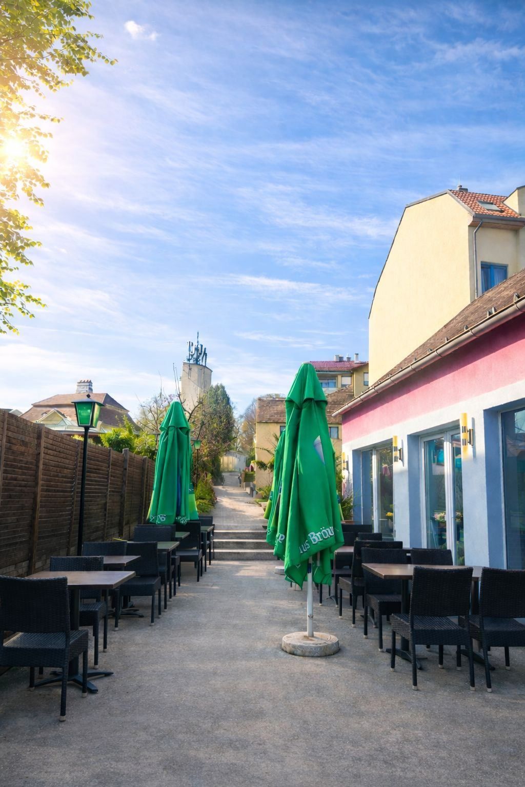 Außenbereich eines Restaurants mit grünen Sonnenschirmen, Holztischen und einer Kulisse aus Gebäuden und Bäumen unter einem klaren blauen Himmel.