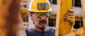 Ein Mann in einem blauen Hemd, gelbem Helm und reflektierenden Sonnenbrillen steht gegen einen gelben Balken.