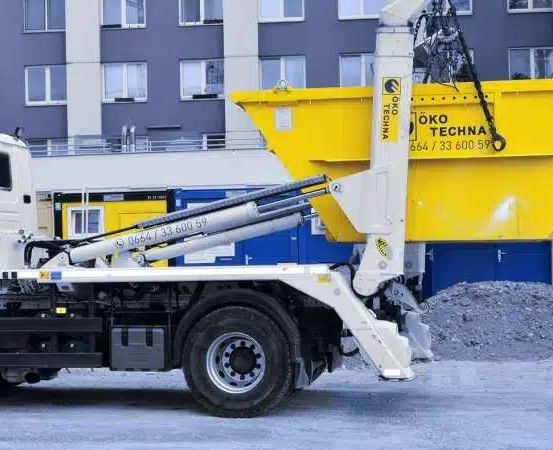 Ein weißer Lkw mit einem gelben Kranaufbau steht vor einem Gebäude mit einem Haufen Kies auf dem Boden.