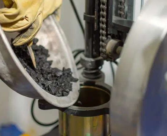 Eine Person mit gelben Handschuhen füllt Kohle in einen Metallbehälter, der mit einer Maschine verbunden ist.