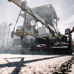 Ein Arbeiter steht neben einer großen Straßenwalze. Die Maschine legt Asphalt auf einer Straße und gibt Dampf ab. Im Hintergrund sind Bäume und Berge zu sehen.