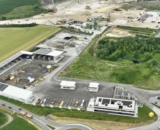 Luftaufnahme einer Baustelle mit mehreren Gebäuden, Solarpanelen und geparkten Fahrzeugen auf einem Parkplatz, umgeben von Grünflächen und einer Straße.