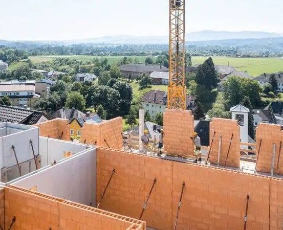 Eine Baustelle mit Arbeitern, Ziegelwänden und einem Kran. Sie hat einen Blick auf die Vorstadt mit Häusern und einem grünen Feld.