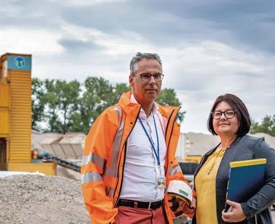 Zwei Personen, ein Mann und eine Frau, stehen vor einer Baustelle. Der Mann trägt eine reflektierende orange Jacke und einen weißen Harthelm. Die Frau, gekleidet in einem gelben Shirt und einer blauen Blazerjacke, hält eine blaue Mappe. Hinter ihnen ist ein gelbes Baufahrzeug zu sehen.