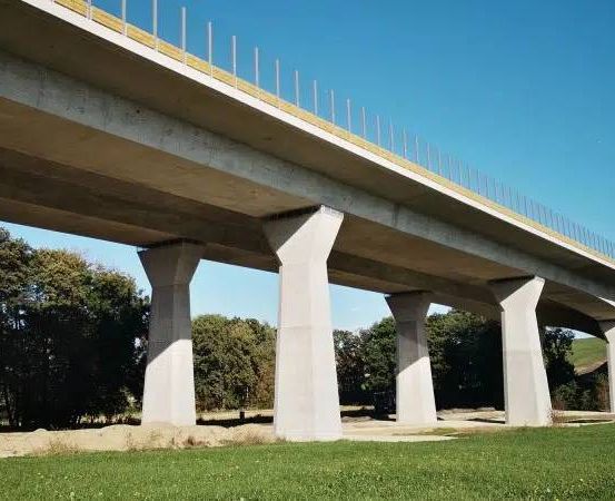 Eine Betonbrücke mit Metallgeländer überspannt ein Tal, gestützt von Säulen und umgeben von Bäumen und Gras unter einem klaren Himmel.