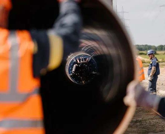 Zwei Arbeiter in orangen Westen inspizieren eine große Rohrleitung, während ein weiterer Arbeiter in blauer Jacke und Helm in der Nähe steht. Die Szene spielt im Freien mit Strommasten im Hintergrund.
