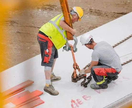 Zwei Arbeiter in Schutzkleidung sind auf einer Baustelle. Einer hält eine Kette und einen Haken, während der andere kniet. Sie arbeiten an einer Betonplatte.