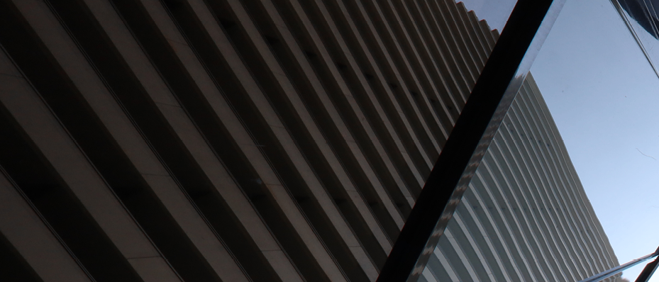Ein geometrisches architektonisches Design mit eckigen Glaspaneelen reflektiert den Himmel. Ein hohes Gebäude mit vertikalen Streifen ist im Hintergrund sichtbar.