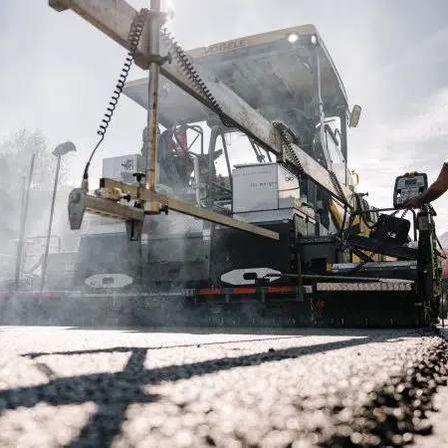 Eine Straßenbaumannschaft verwendet eine Asphaltiermaschine, um Asphalt auf einer Straße zu verlegen, mit Bäumen und Bergen im Hintergrund.