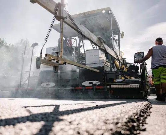 Eine Straßenbaumannschaft verwendet eine Asphaltiermaschine, um Asphalt auf einer Straße zu verlegen, mit Bäumen und Bergen im Hintergrund.