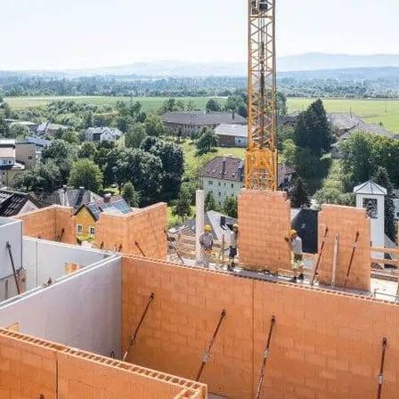 Ein Luftbild einer Baustelle mit im Bau befindlichen Ziegelwänden, einem Kran und einer Stadtlandschaft im Hintergrund.