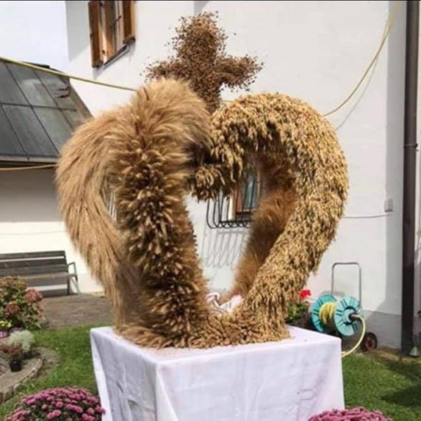 Eine herzförmige Skulptur aus Weizenhalmen steht auf einem weißen Tisch im Garten, mit einem Gebäude und einer Bank im Hintergrund.