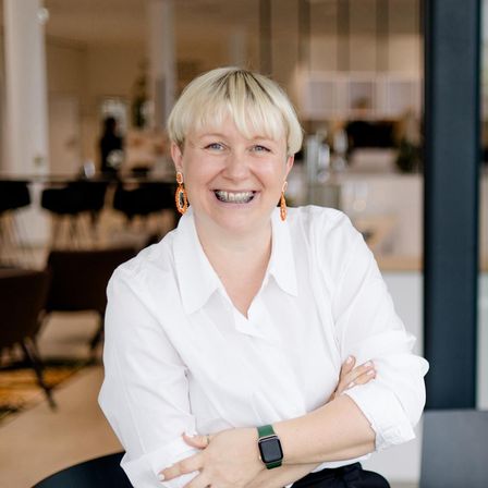 Eine Frau mit kurzen blonden Haaren, die ein weißes Hemd und orangefarbene Ohrringe trägt, sitzt mit verschränkten Armen in einem Büro.