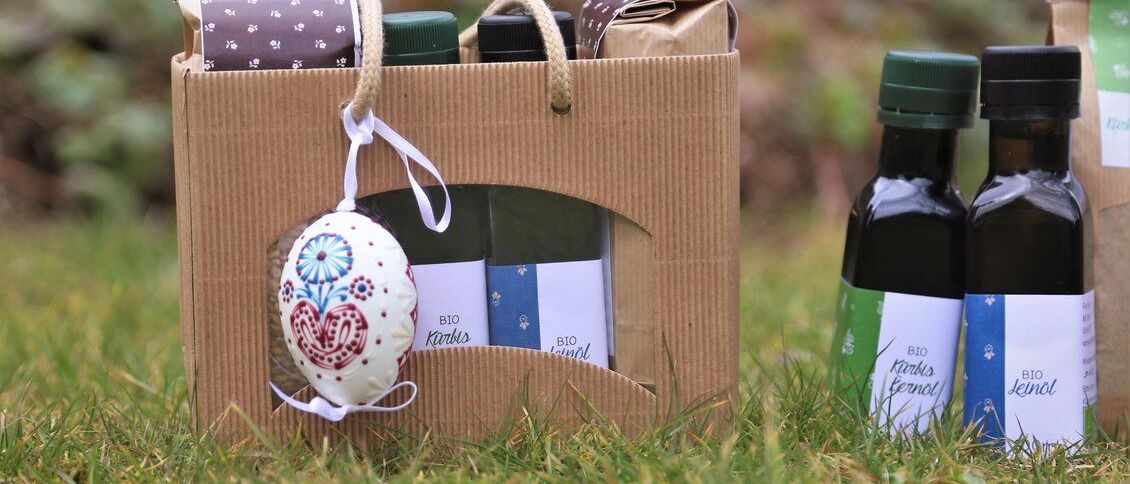 Ein brauner Papierbeutel enthält drei Flaschen Olivenöl, eine Flasche mit blauem Etikett und ein Osterei. Draußen befindet sich eine weitere Flasche Olivenöl mit grünem Etikett.