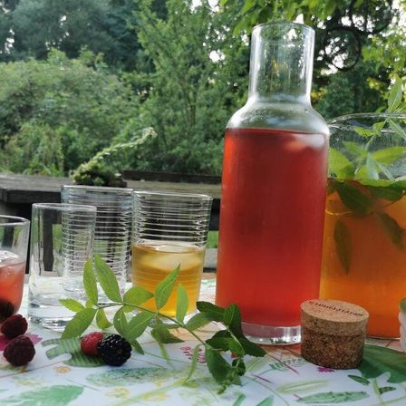 Ein Tisch mit Getränken, Gläsern, Beeren, Blättern und einem Korken im Garten. Die Getränke sind in Krügen und Gläsern. Die Blätter sind Minze.