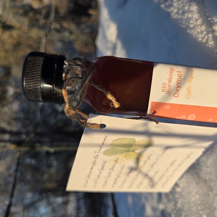 Eine Flasche Bio-Schafgarben-Syrup, in eine Schnur gewickelt und begleitet von einem Etikett mit einem Blumendesign, ruht auf einer verschneiten Oberfläche.