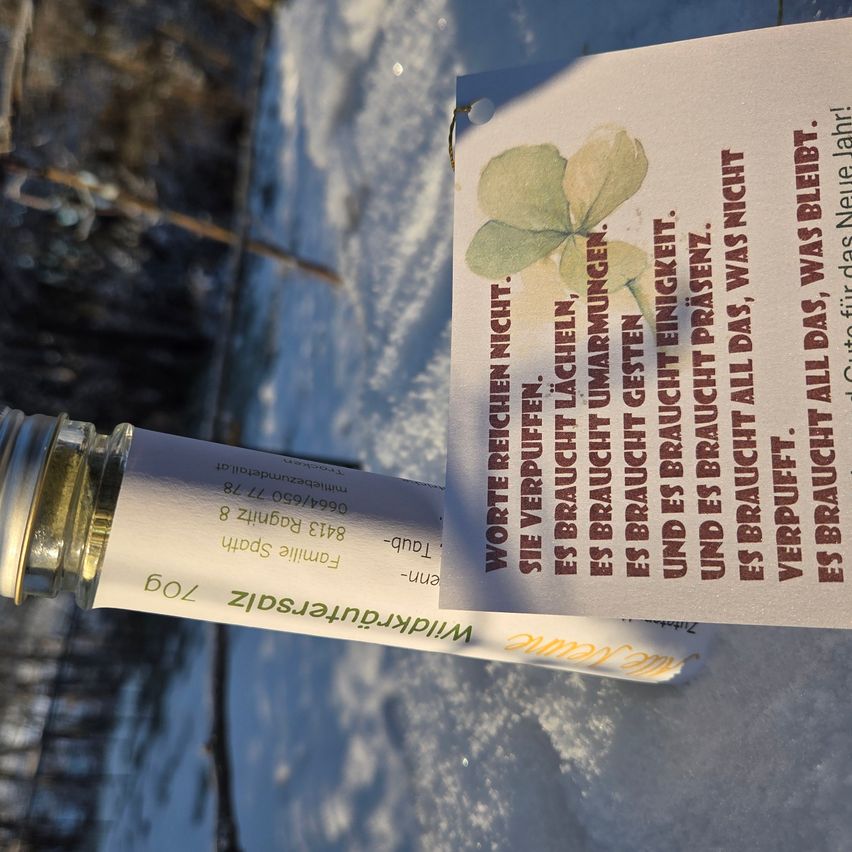 Ein kleiner, weißer Behälter mit der Aufschrift 'Microgreens' befindet sich neben einem Stück Papier mit einer Illustration eines grünen Kleeblatts und deutschem Text. Der Hintergrund ist verschneit und unscharf.