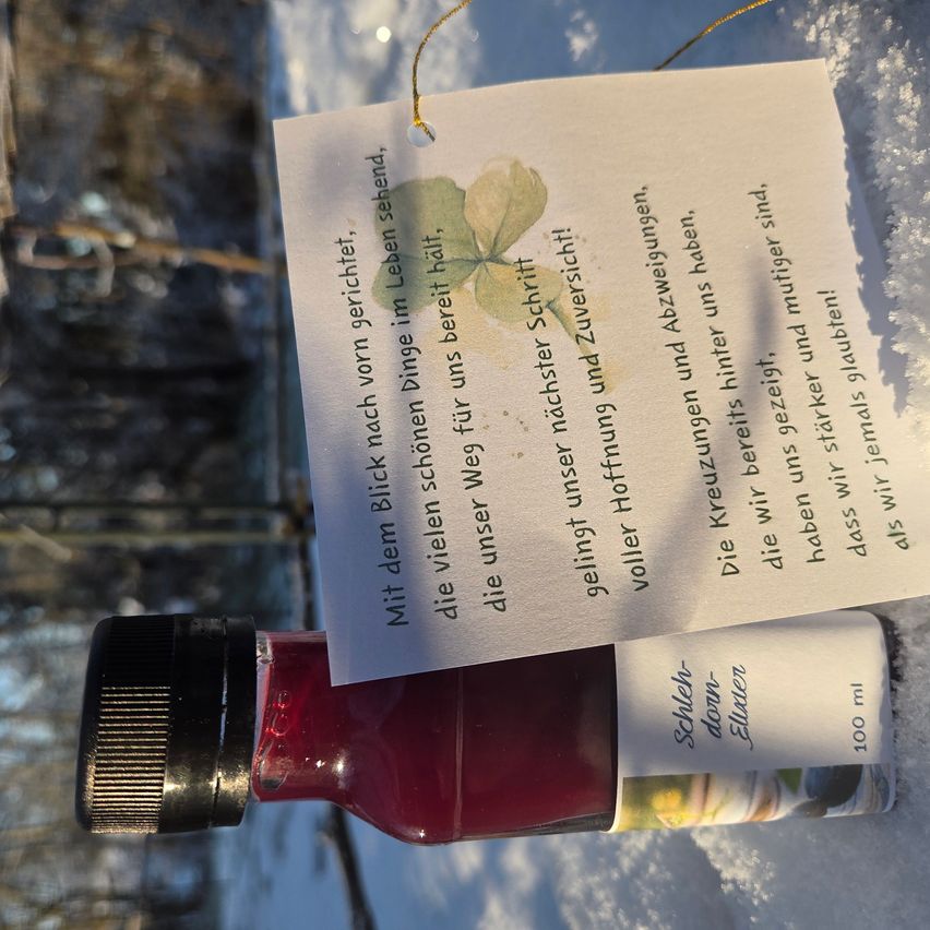 Eine rote Flasche mit schwarzem Deckel liegt auf einer verschneiten Oberfläche. Ein Etikett ist befestigt und zeigt Text und eine Illustration eines Blattes.