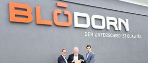 Drei Männer stehen vor einem Gebäude mit dem Schild 'BLCDOR'. Der Mann in der Mitte hält ein Zertifikat. Sie sind alle formell gekleidet.