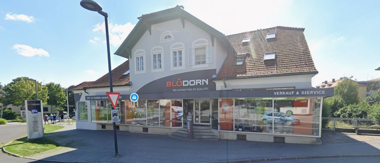 Ein Ladenlokal mit dem Wort Blodorn in fetten Buchstaben auf einem Vordach. Der Laden hat Glastüren und Fenster mit der Reflexion eines Autos. Die Straße hat eine Straßenlaterne und ein Schild.