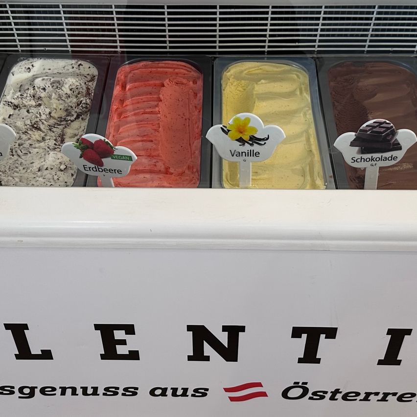 Eine Eiscreme-Vitrine zeigt vier Geschmacksrichtungen: Schokolade, Erdbeere, Vanille und eine weitere Geschmacksrichtung. Die Anzeige ist mit 'Lenti' und 'Genuss aus Osterreich' beschriftet.