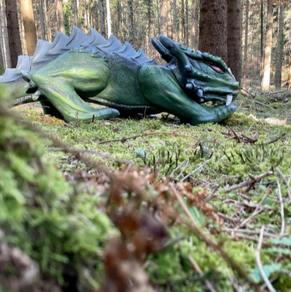 Eine grüne Drachenfigur liegt auf moosbedecktem Boden in einem Wald, ihre Augen leuchten rot. Die Baumstämme sind hoch und dick, und der Waldboden ist mit Blättern und Moos bedeckt.