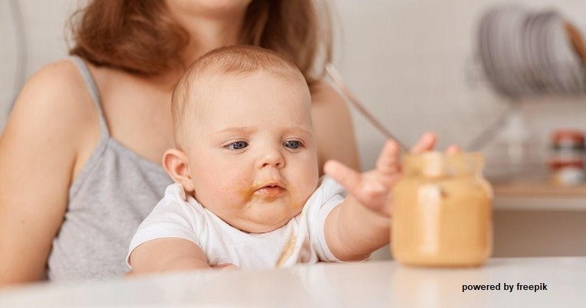 Ein Baby in einem weißen T-Shirt sitzt mit einem Glas Erdnussbutter am Tisch und streckt mit schmutzigem Gesicht die Hand aus.