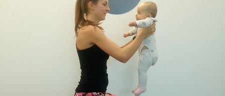 Eine Frau in einem schwarzen Tanktop hält ein Baby in den Armen. Das Baby schaut zu ihr, während beide drinnen an einer weißen Wand stehen.