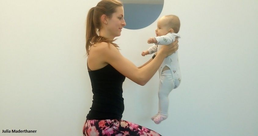Eine Frau in einem schwarzen Tanktop hält ein Baby in den Armen. Das Baby schaut zu ihr, während beide drinnen an einer weißen Wand stehen.