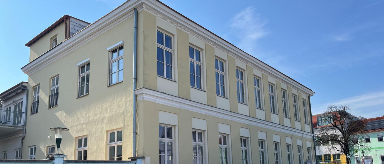 Ein gelbes Gebäude mit vielen Fenstern ist mit einem Schild für Mayer & Co, ein Recycling- und Abrissunternehmen, eingezäunt. Ein Bärenbild ziert den Zaun.