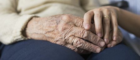 Eine ältere Frau sitzt auf einem Stuhl und hält die Hand einer Frau in einem blauen Hemd. Die ältere Frau trägt einen beigefarbenen Pullover.