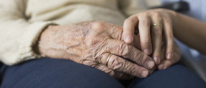 Eine ältere Frau sitzt auf einem Stuhl und hält die Hand einer Frau in einem blauen Hemd. Die ältere Frau trägt einen beigefarbenen Pullover.